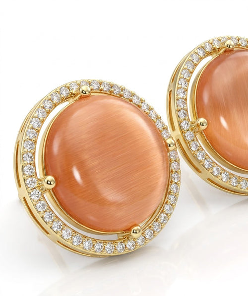 A pair of gold round stud earrings featuring large peach cabochon gemstones surrounded by a halo of small clear stones, set against a clean white background.