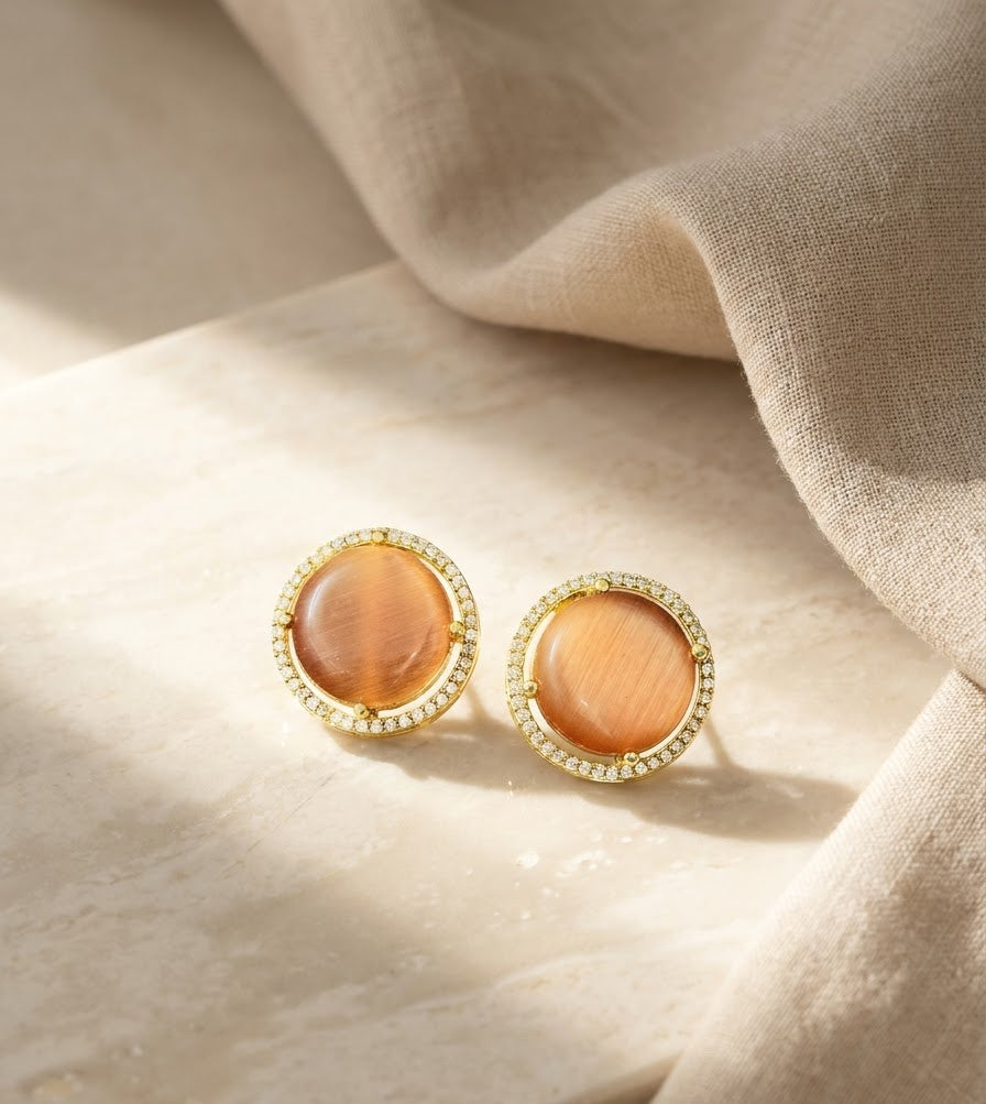 A pair of gold-toned stud earrings with oval amber-orange stone centers and a halo of small clear stones, styled on a light stone surface with draped beige fabric.