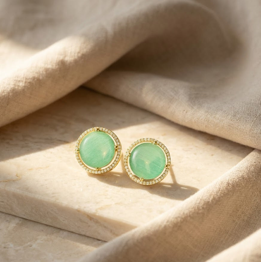 A pair of gold round stud earrings with light aqua blue circle centre, accented with small clear stones, resting on a beige stone slab with draped linen fabric in soft sunlight.