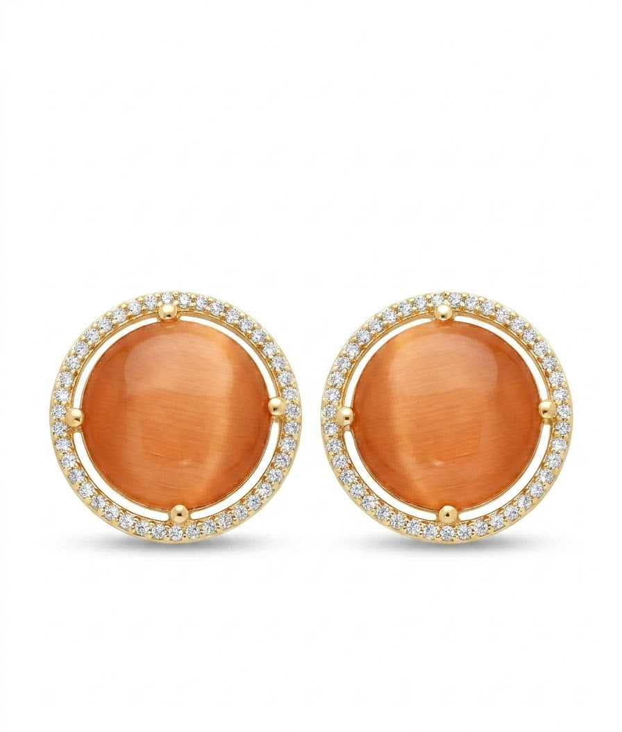 A pair of gold round stud earrings featuring large orange cat's eye cabochons surrounded by a halo of small clear pavé stones, set against a plain white background.