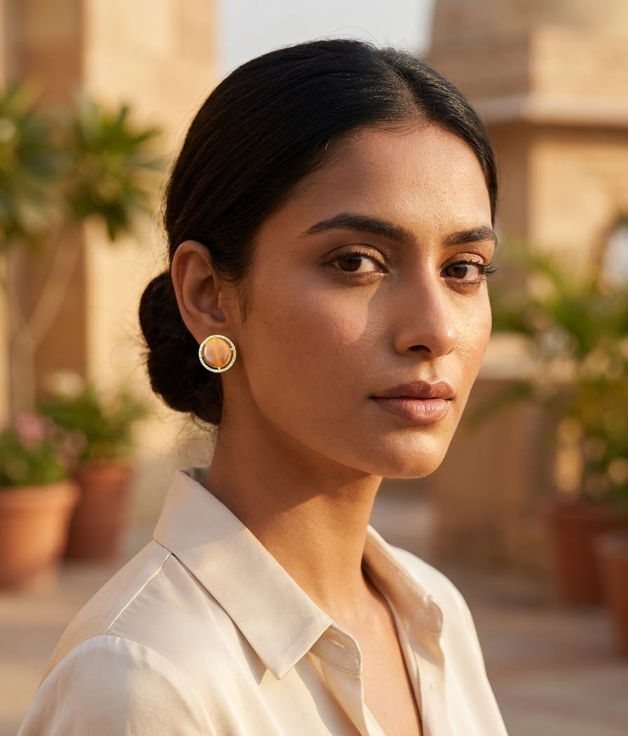 A woman with dark hair tied back wears a small gold hoop earring with a circular beige inlay, photographed in warm outdoor light against a softly blurred architectural background with potted plants.