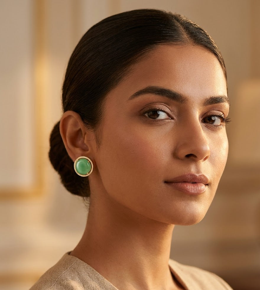 A woman with her hair in a low bun wears a round aqua blue earring with a gold-toned bezel in a warm, softly lit indoor setting.
